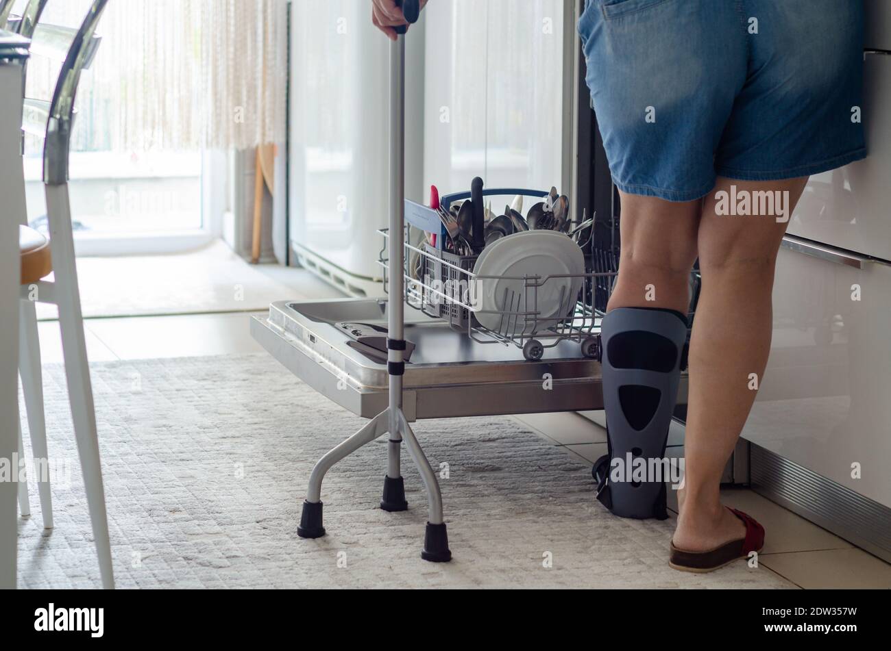 The woman with a orthotics on her foot is emptying the dishwasher in the kitchen Stock Photo