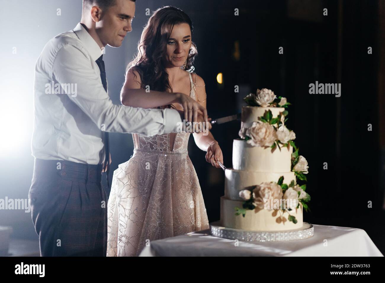 Concentrated newlyweds cutting three-tiered wedding cake decorated with flowers. Wedding party. Happy bride and groom Stock Photo