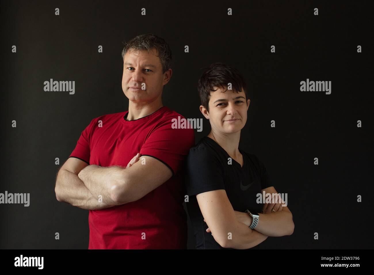Caucasian athletic personal trainer couple dressed in red and in black posing back on back with crossed arms in front of a black background. Stock Photo