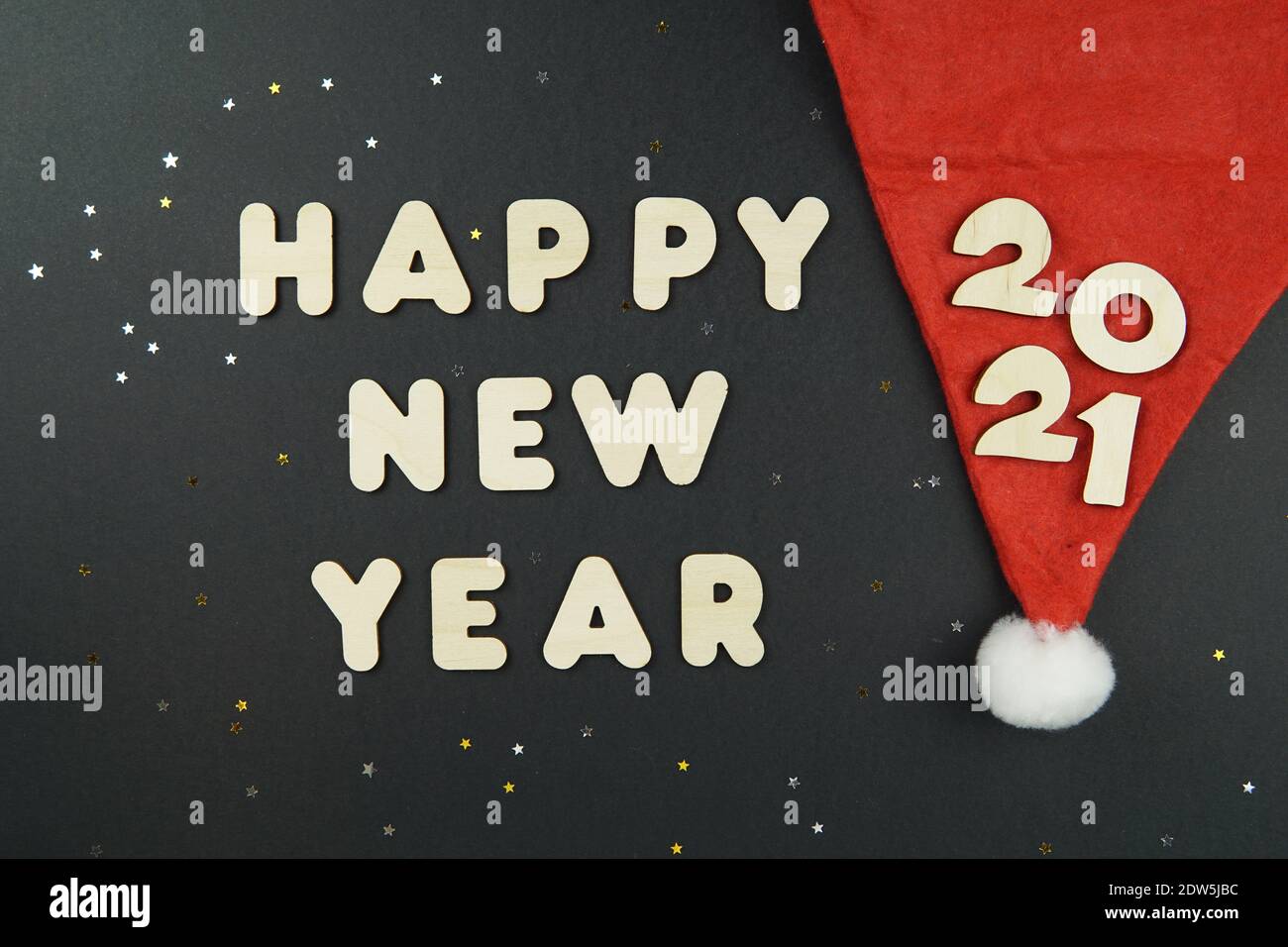 Wooden numbers 2021 on a red Santa hat and words HAPPY NEW YEAR on a black background with yellow and gray glitter stars. Stock Photo