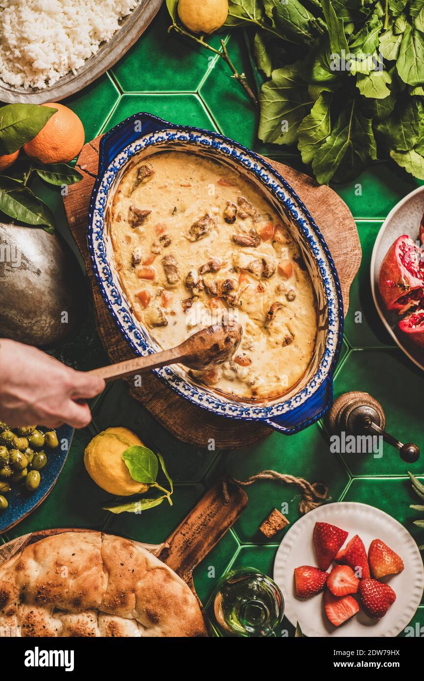 Turkish style family dinner. Flat-lay of female hands stirring Turkish lamb in yogurt sauce over table with rice pilav, fresh arugula and strawberry s Stock Photo