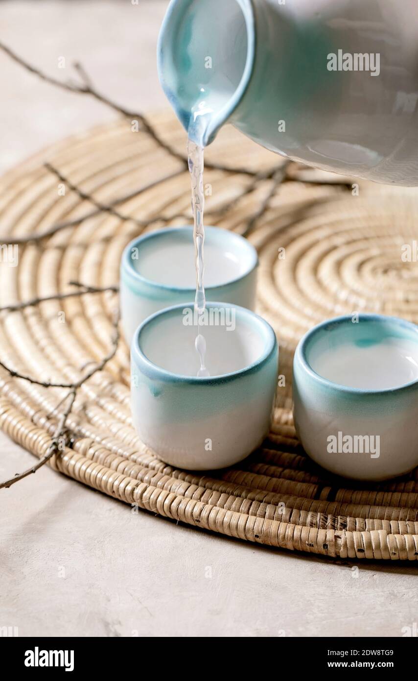 Sake ceramic set for traditional japanese alcohol drink rice wine sake pouring from pitcher in three cups, standing on straw napkin with dry branches Stock Photo