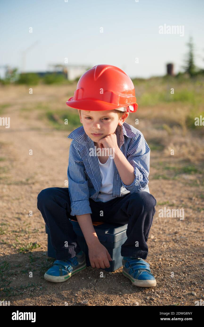 upset little kid in orange helmet sitting on road to factory industrial plant being Stock Photo