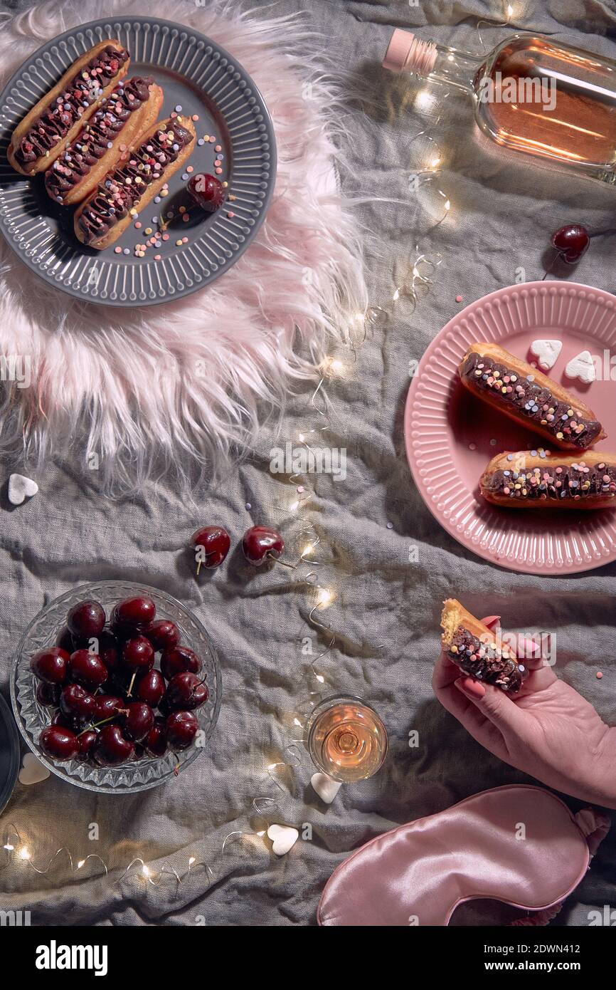 Happy Valentine celebration Trendy flat lay with assortment of yummy eclairs or brewing cakes and cherry. Grey linen background with pink fur and Stock Photo