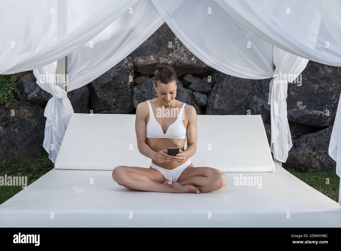 Glad female wearing white bikini sitting with crossed legs on canopy bed  and surfing Internet on mobile phone while relaxing during summer holiday  Stock Photo - Alamy