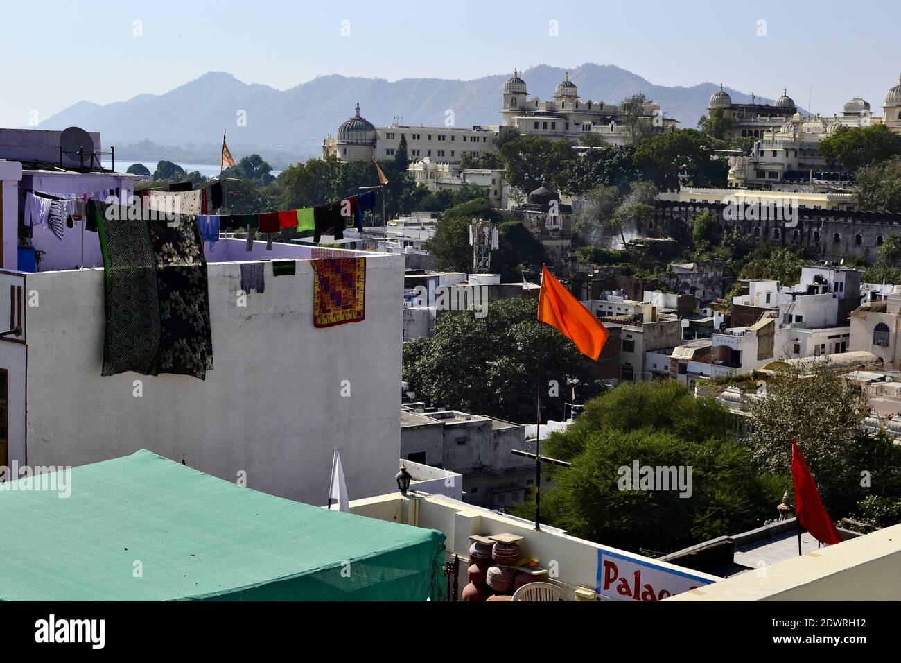 Morning rooftop view of Udaipur City Palace and Pichola lake. Mountain range and blue sky on the background. Orange bright  waving flag. Rajasthan Stock Photo