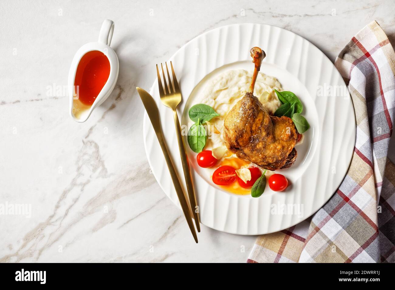 Roasted duck leg - duck confit with parsnip puree and orange sauce cherry tomatoes, fresh spinach leaves served on a white plate with golden cutlery o Stock Photo