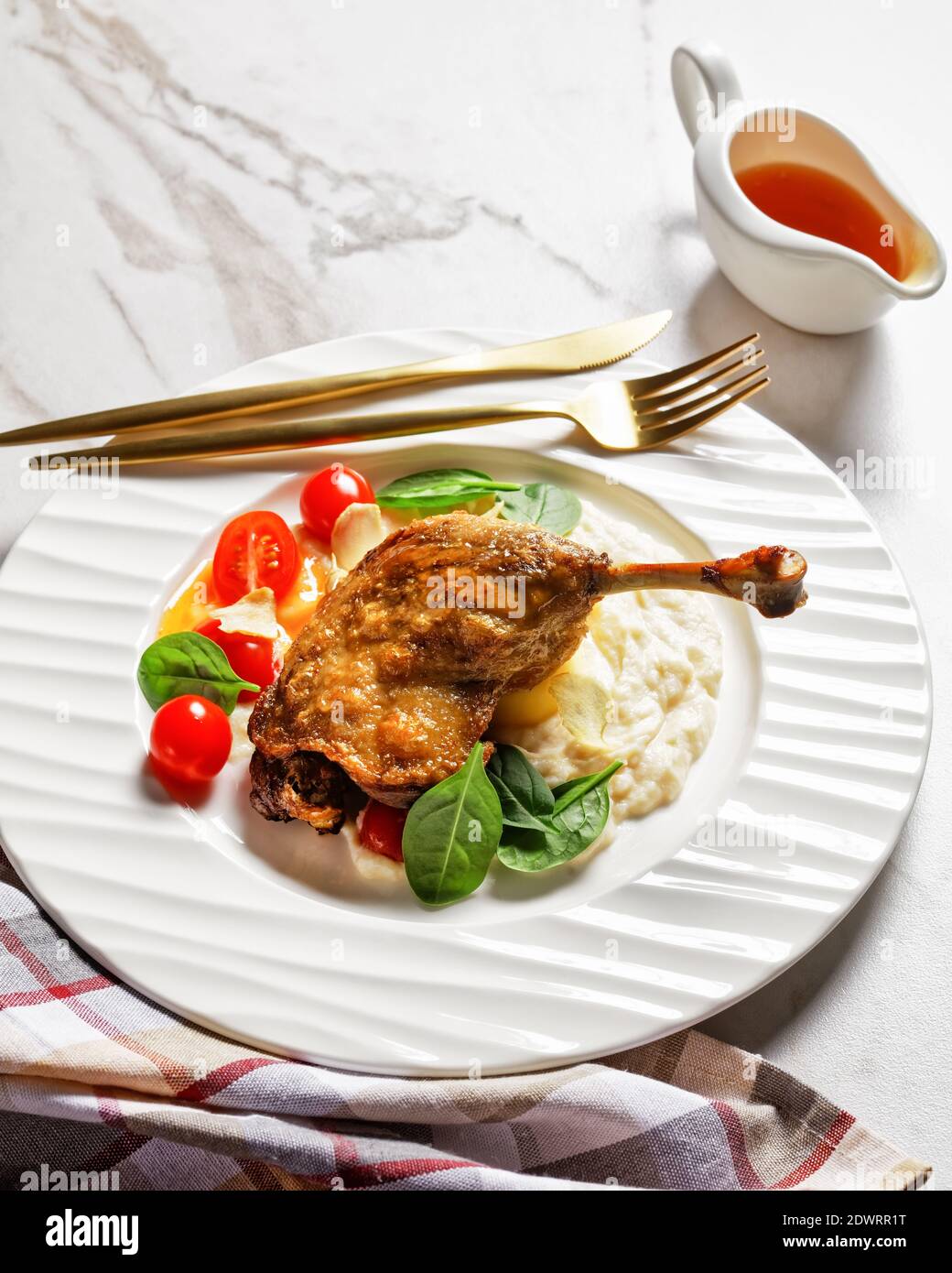 Roasted duck leg - duck confit with parsnip puree and orange sauce cherry tomatoes, fresh spinach leaves served on a white plate with golden cutlery o Stock Photo