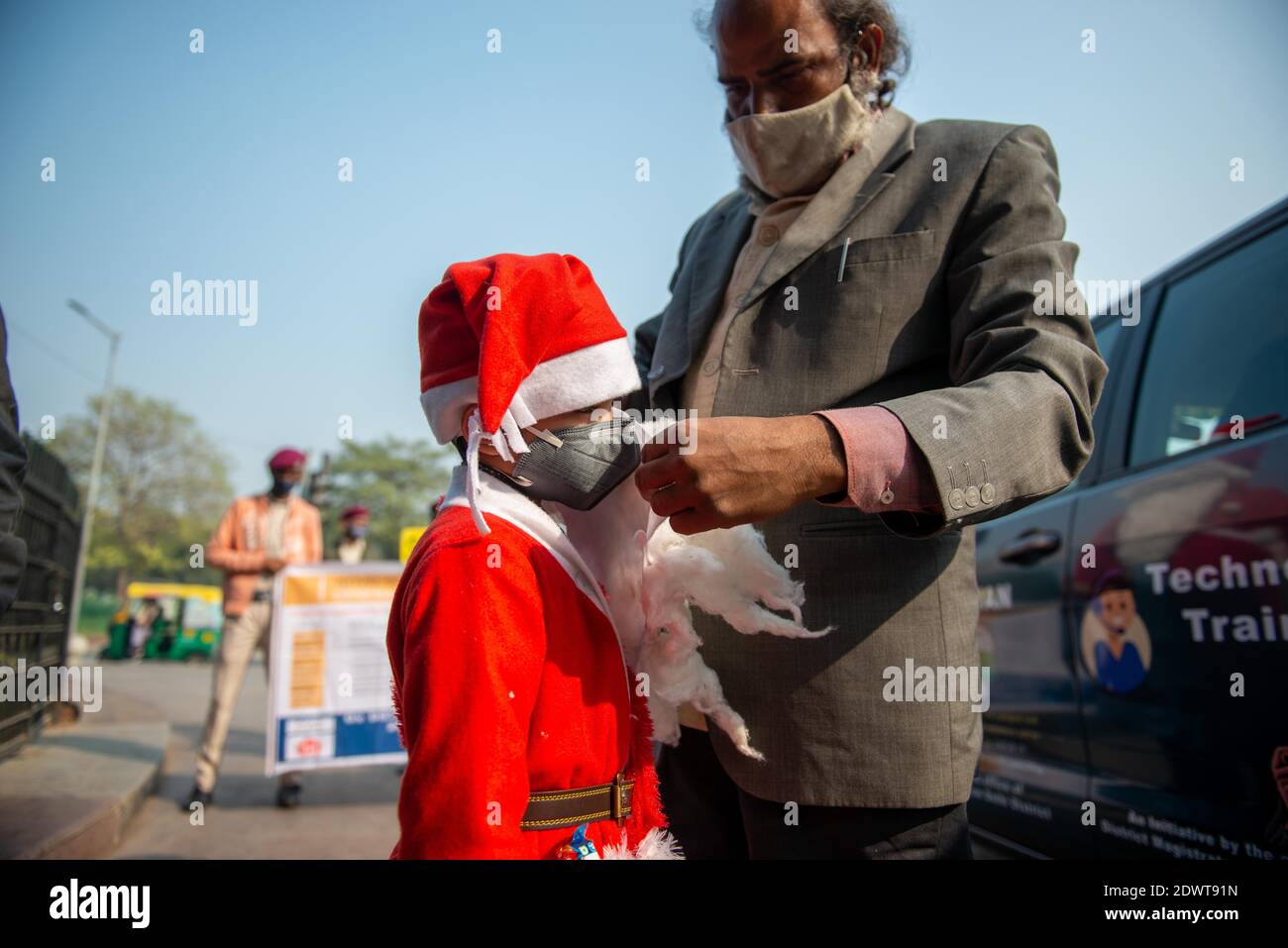 New Delhi, India. 23rd Dec, 2020. A man assisting a kid dressed up as Santa Claus to wear a face mask during the awareness campaign.The awareness campaign took place in Connaught Place, Delhi organized by the Office of District Magistrate to make people aware of Coronavirus pandemic precautionary measures. An artist and a boy attired like Santa Claus distributes masks among daily commuters. Credit: SOPA Images Limited/Alamy Live News Stock Photo