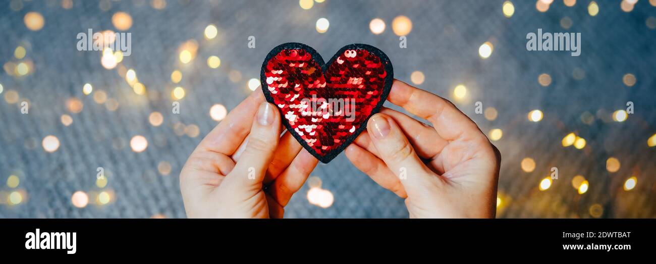 Red sequins heart in woman's hands on gray background with bokeh lights. Valentine's day and love concept. Web banner. Stock Photo