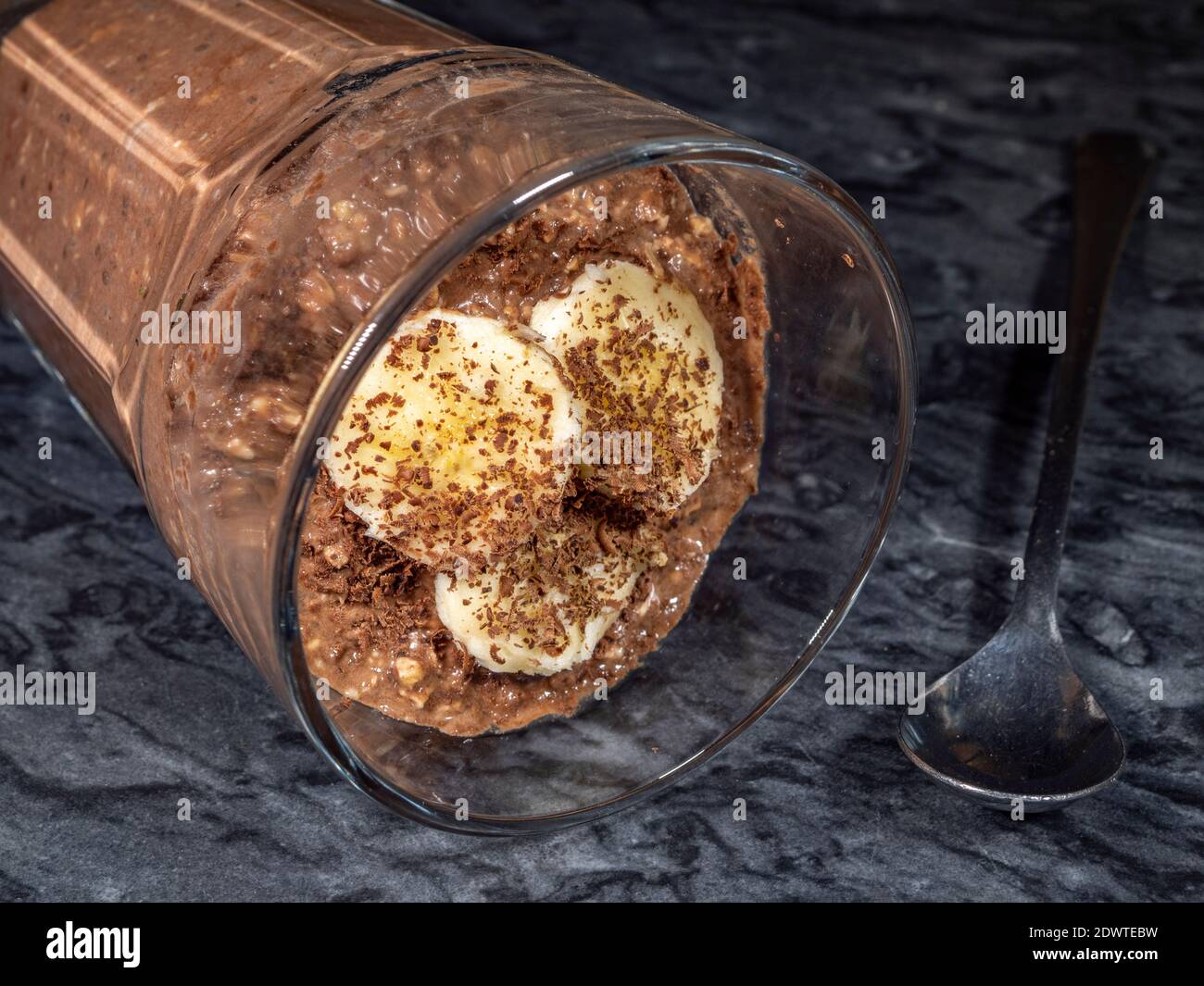 Closeup POV shot of a glass tumbler on its side, full of thick textured, chocolate overnight oats with banana topping, which has set (breakfast food). Stock Photo