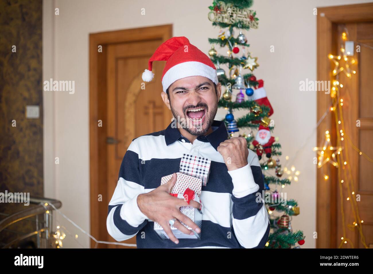 Cheerful Indian Man Wearing Santa hat Holding Christmas Gift Boxes Or Presents Enjoys Holiday Season At Home, Extreme Happiness,Screams,Crazy Emotions Stock Photo
