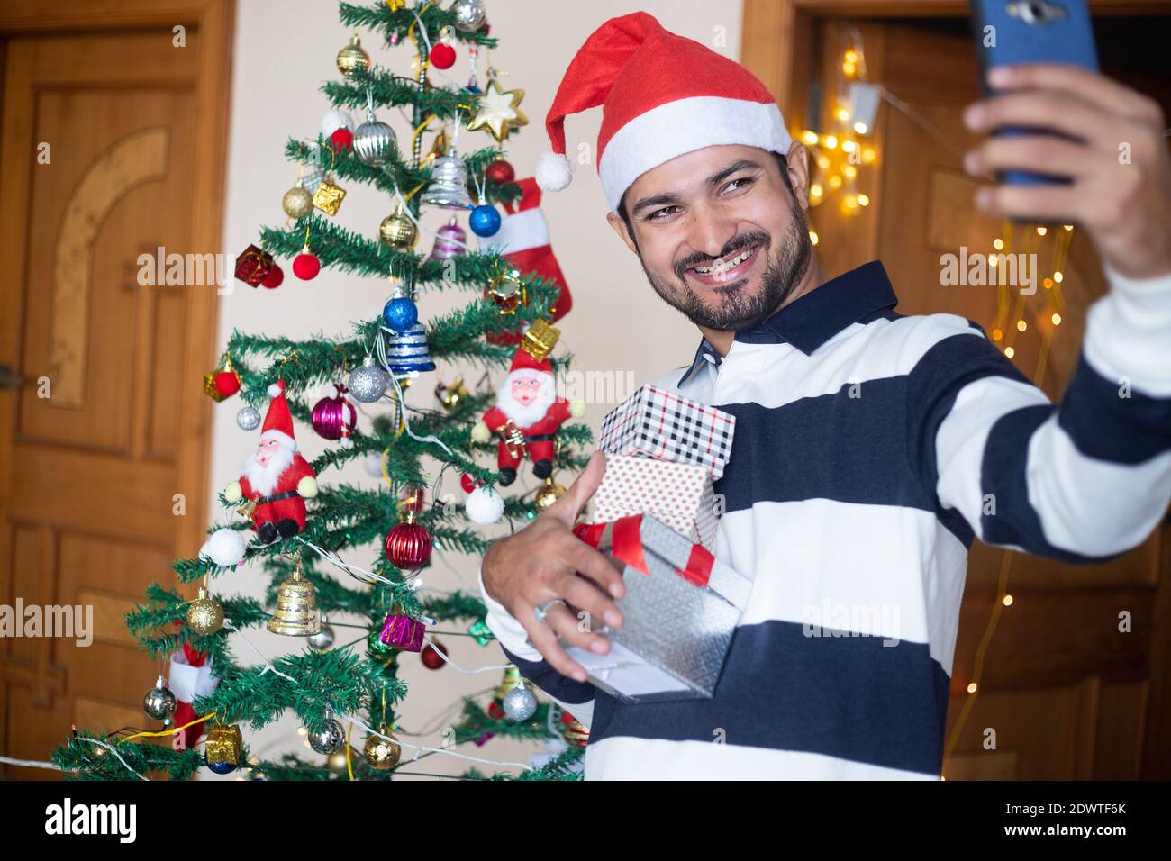 Happy Indian Man Wearing Santa hat Talking Selfie Or Video Call Over Smart Phone Holding Christmas Gift Boxes Or Presents Enjoys Holiday Season At Hom Stock Photo