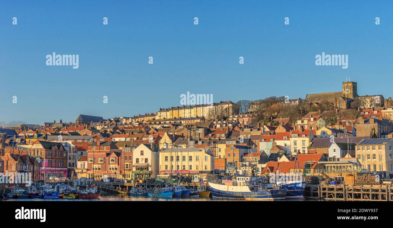 A low winter sun falls on the buildings of a town that is set on a hillside leading up from the  harbour below. A clear blue sky is above. Stock Photo