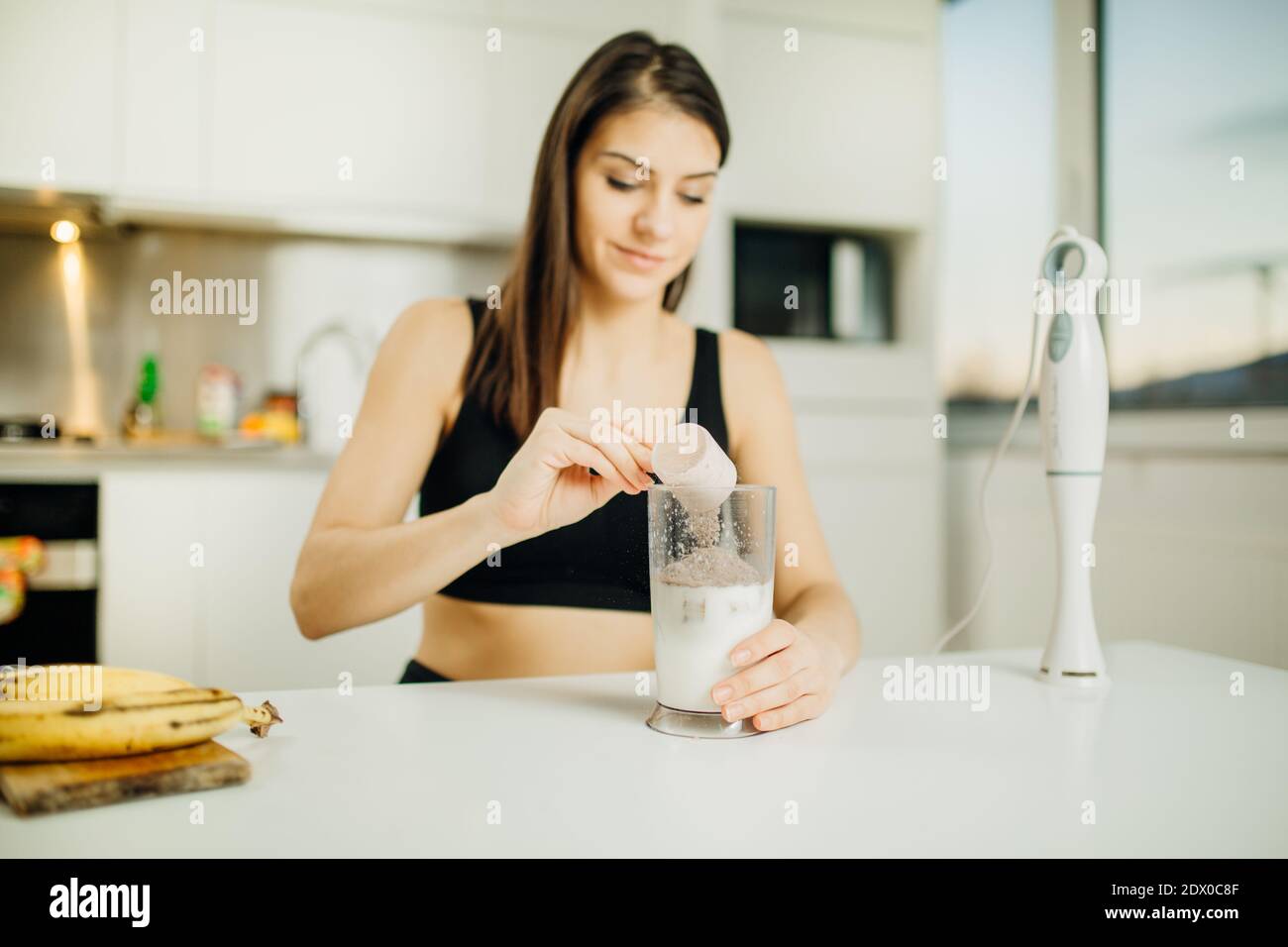 Woman with immersion blender making banana chocolate protein powder milkshake smoothie.Adding a scoop of low carb whey protein mix to shake after a ho Stock Photo