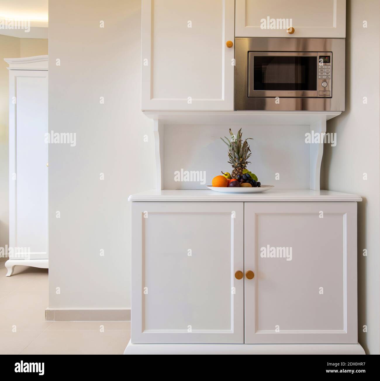 Front view of mini kitchen cupboard module cabinet with microwave and fruits plate. Classic style interior of white room with empty copy space wall Stock Photo