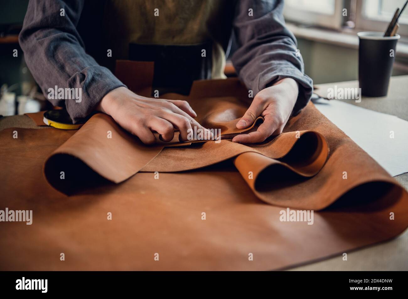 A young apprentice in a boot workshop prepares leather for further use on a large table. Stock Photo