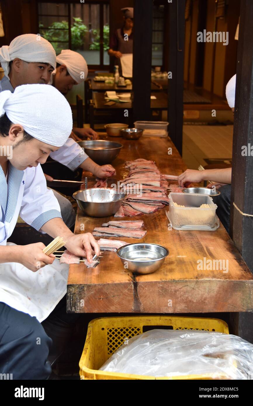 Young Japanese chefs, wearing tenugui headwear, preparing raw fresh eels on the skewers for grilling in front of the restaurant in Narita, Japan. Stock Photo