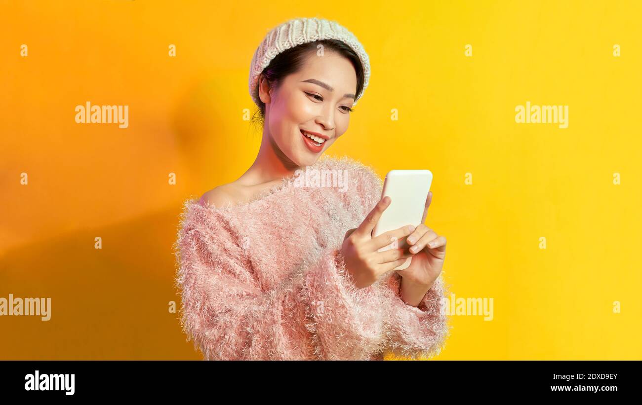 Pretty lady looks at telephone screen, chatting messages, wear warm casual hat, posing isolated over yellow background, Stock Photo