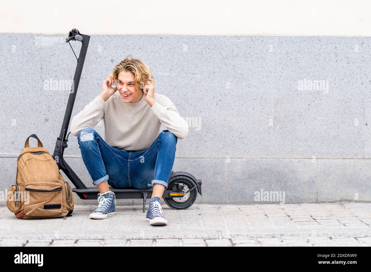 Smiling man with backpack wearing headphones while sitting on electric push scooter against wall Stock Photo