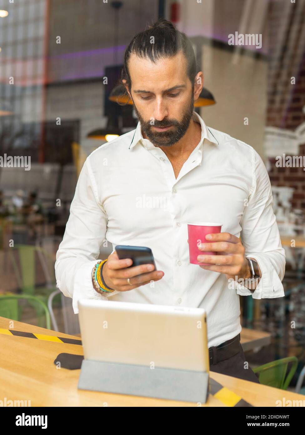 Candid portrait of bearded businessman drinking coffee and using phone inside cafe Stock Photo