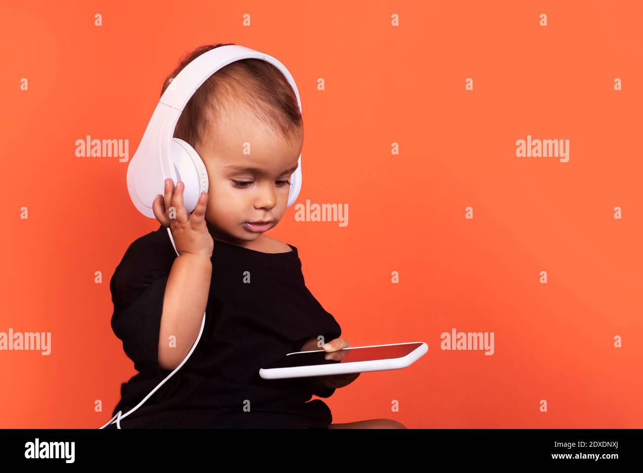 Cute baby girl wearing headphones using mobile phone while sitting against orange background Stock Photo