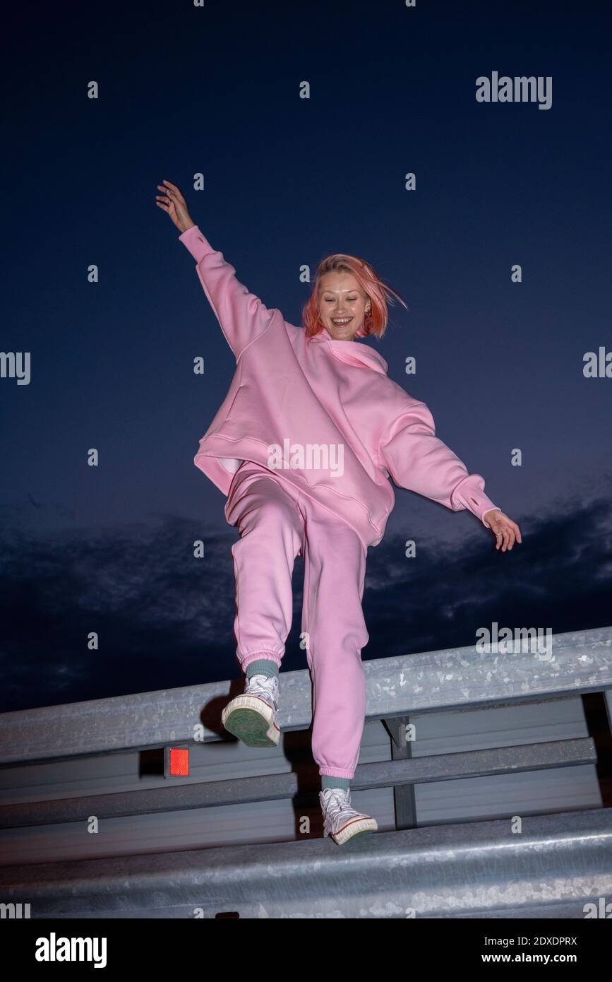 Young woman with pink hair wearing pink track suit standing on one leg on road barrier at night Stock Photo