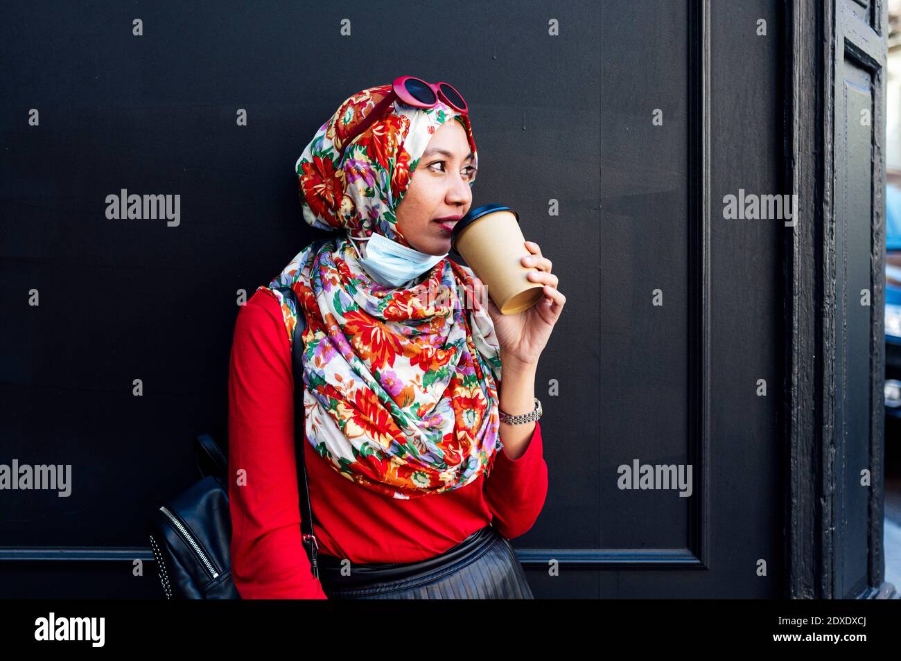 Muslim woman while drinking coffee against black wall during COVID-19 Stock Photo
