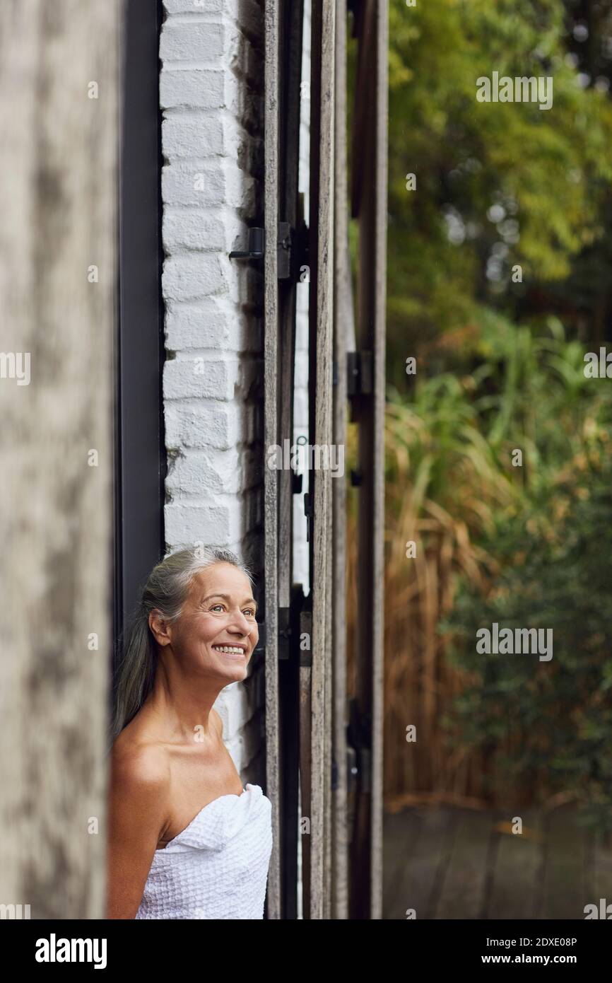Happy mature woman in towel looking away while standing at doorway Stock Photo