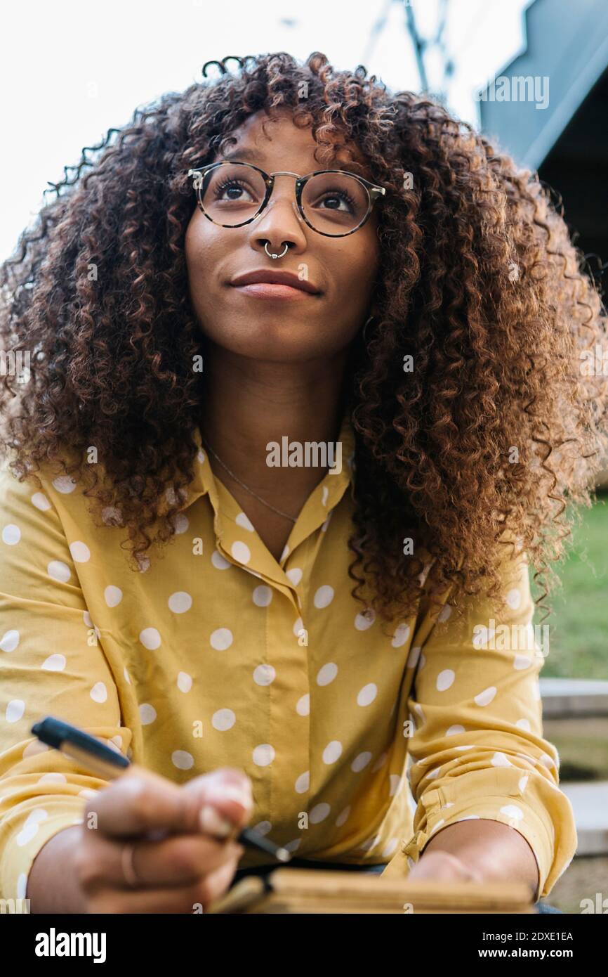 Young thoughtful woman with book looking away while sitting outdoors Stock Photo