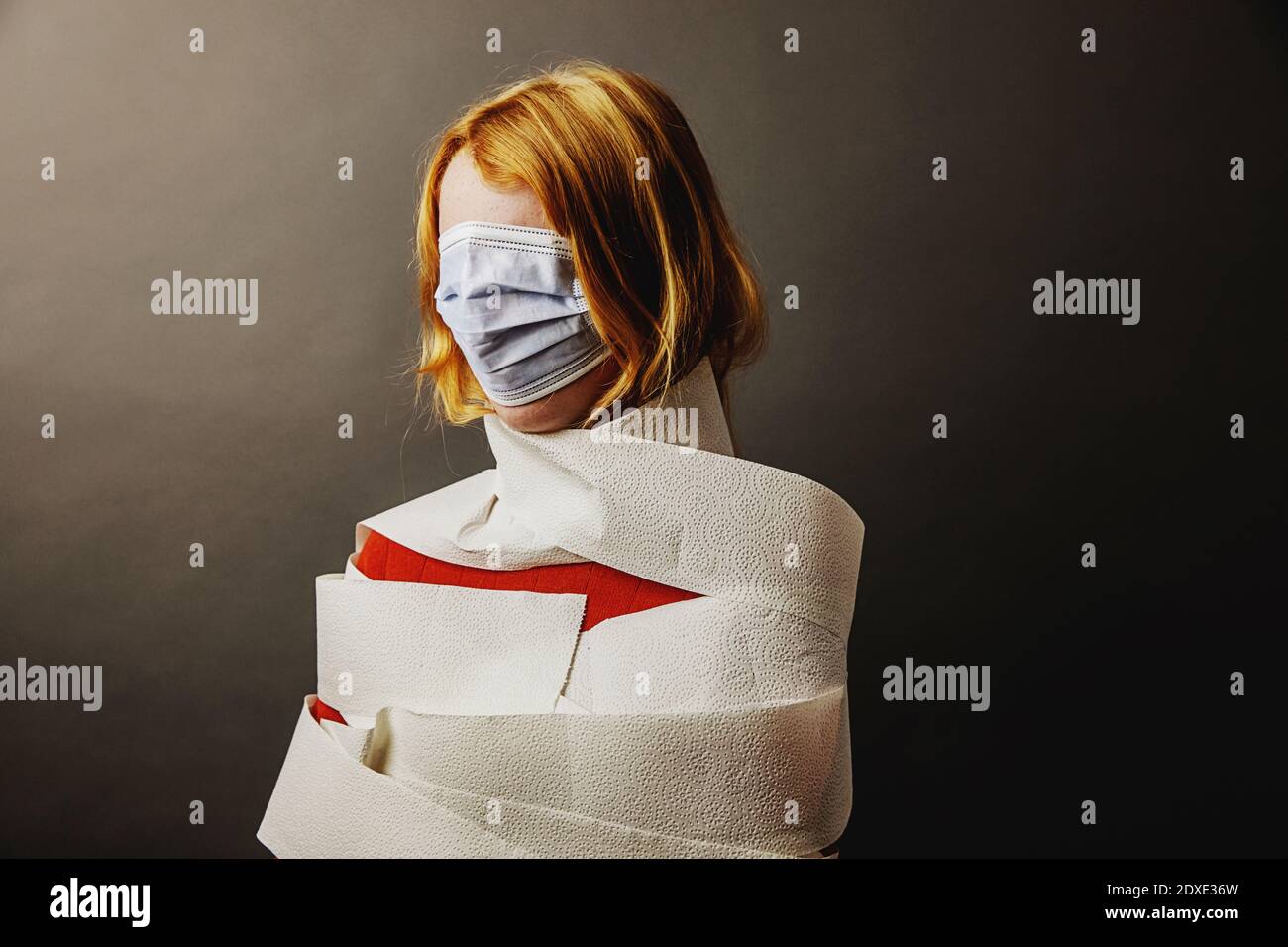 Teenage girl wrapped in toilet paper and face covered with protective face mask standing against gray background Stock Photo