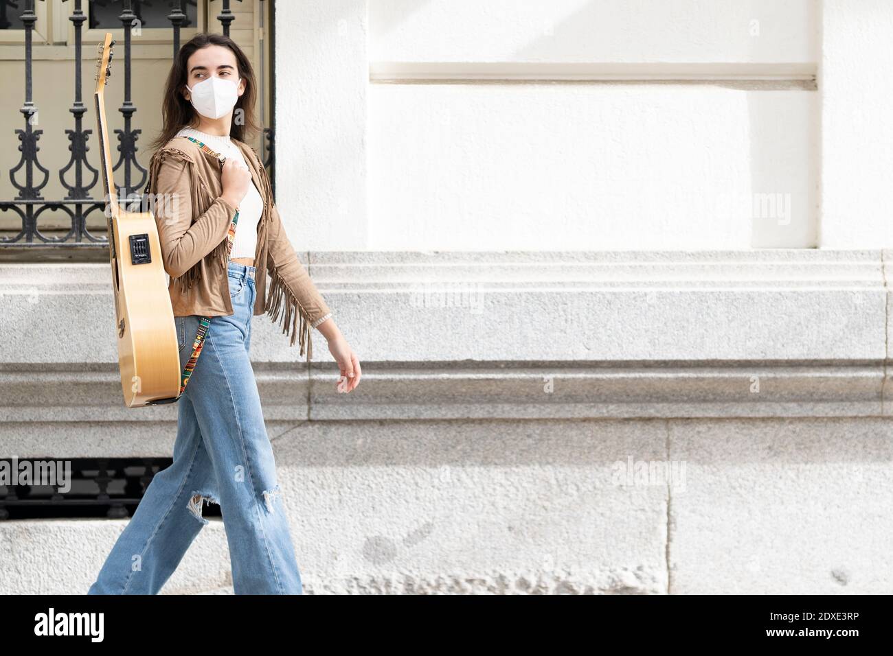 Female musician walking with guitar by building in city during pandemic Stock Photo