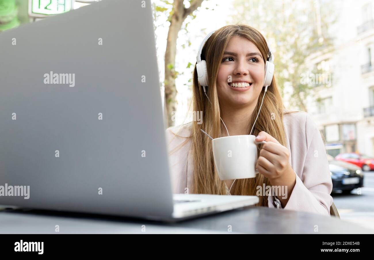 Woman wearing headphones drinking coffee while sitting with laptop at sidewalk cafe Stock Photo