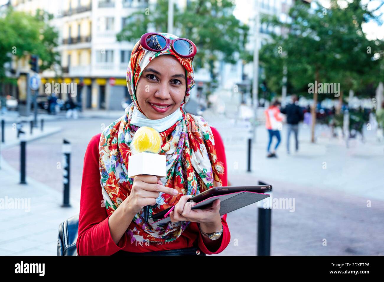 Smiling Muslim female journalist holding digital tablet while reporting in city during pandemic Stock Photo