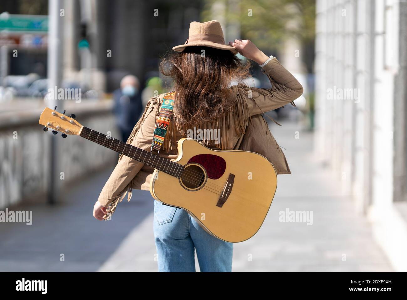 Young woman with guitar wearing hat walking on footpath in city Stock Photo