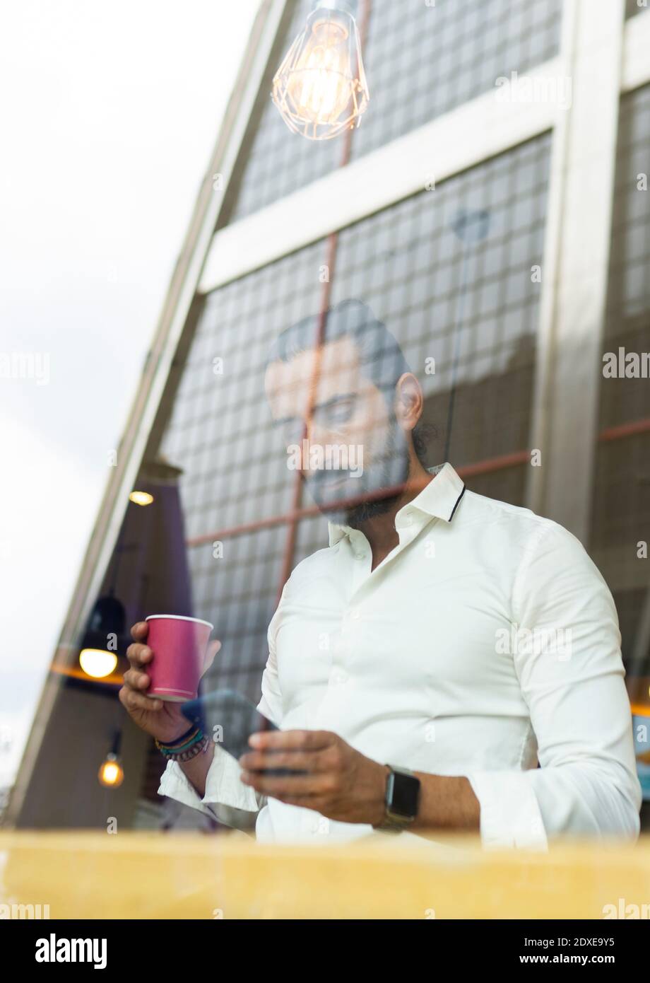 Candid portrait of bearded businessman drinking coffee and using phone inside cafe Stock Photo