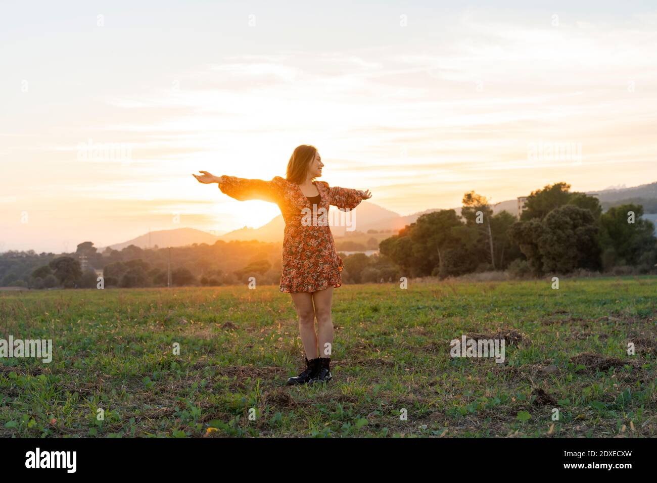 Young woman in sundress with arms outstretched against sky during sunset Stock Photo