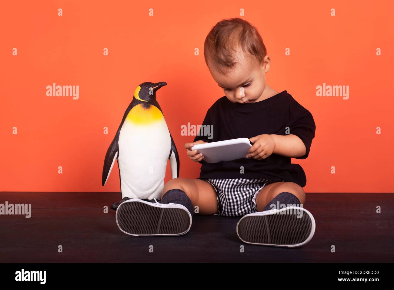 Baby girl ignoring toy while using mobile phone sitting against orange background Stock Photo