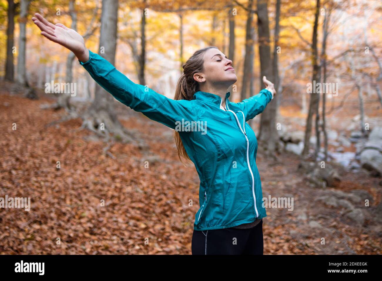 Carefree sportswoman with eyes closed and arms outstretched standing in forest Stock Photo