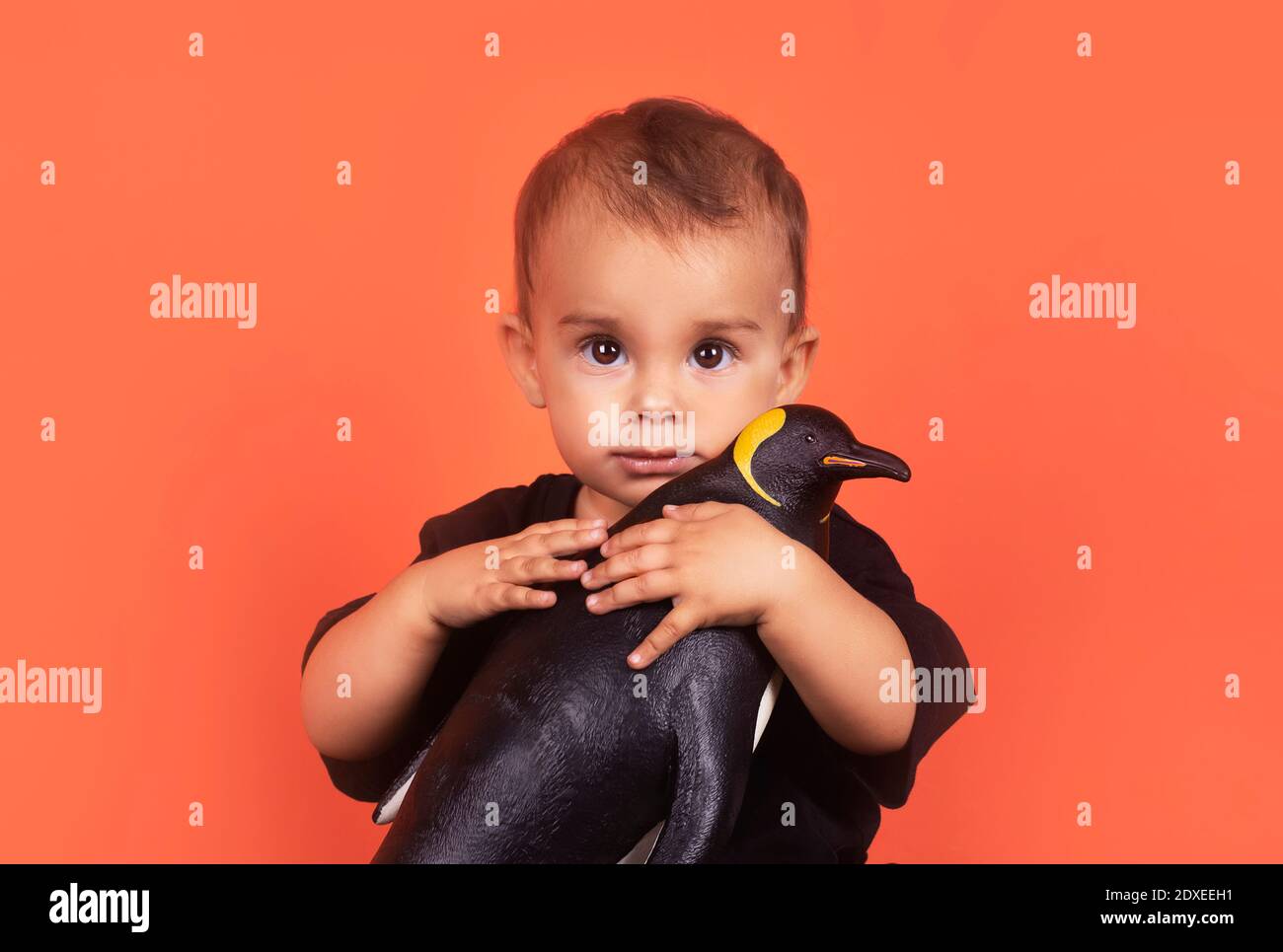 Cute baby girl hugging penguin toy while sitting against orange background Stock Photo