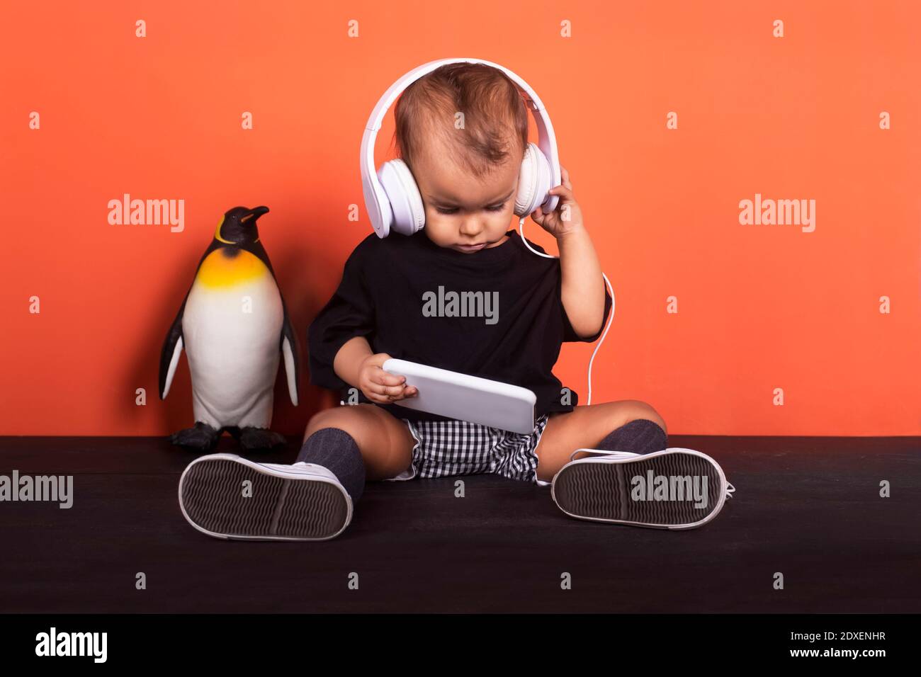 Baby girl wearing headphones using mobile phone while sitting by toy penguin against orange background Stock Photo