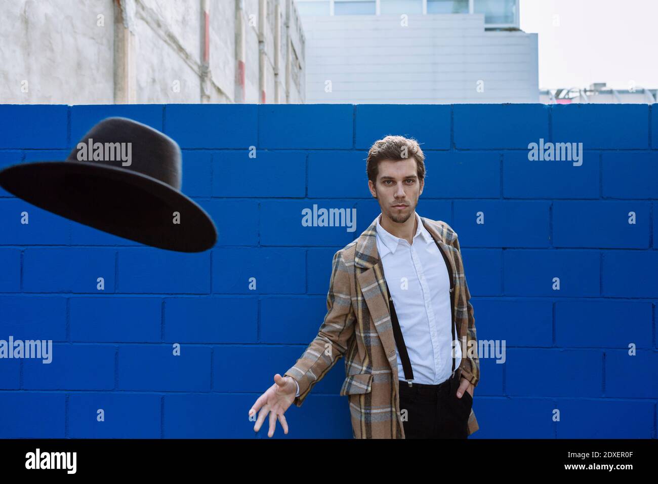 Stylish man wearing checked jacket throwing black hat against blue wall Stock Photo