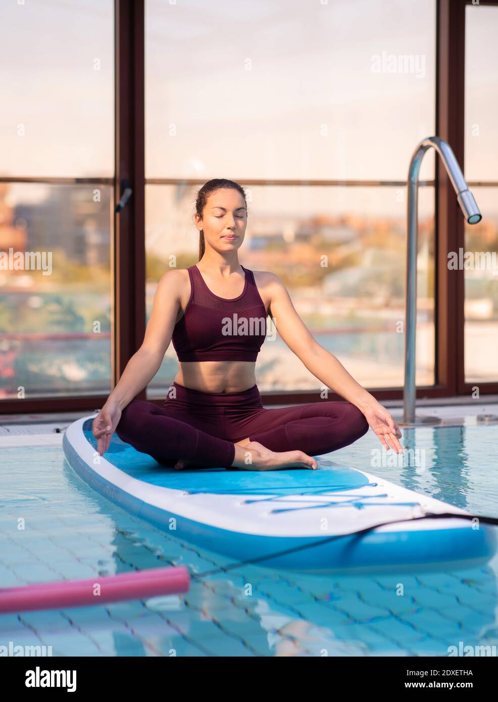 Mid adult female instructor meditating on paddleboard over swimming pool Stock Photo