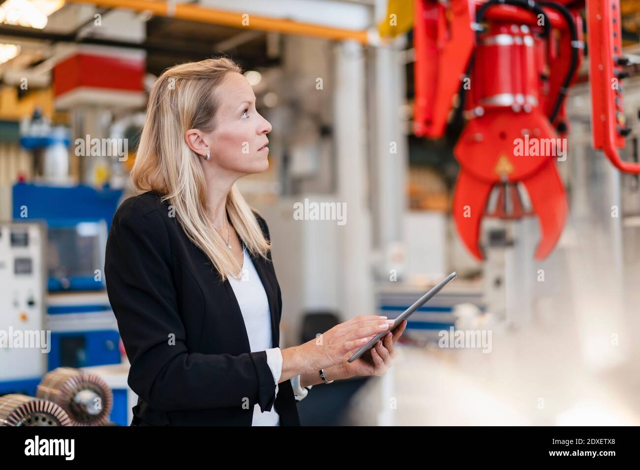 Blond female entrepreneur analyzing claw while holding digital tablet standing in factory Stock Photo