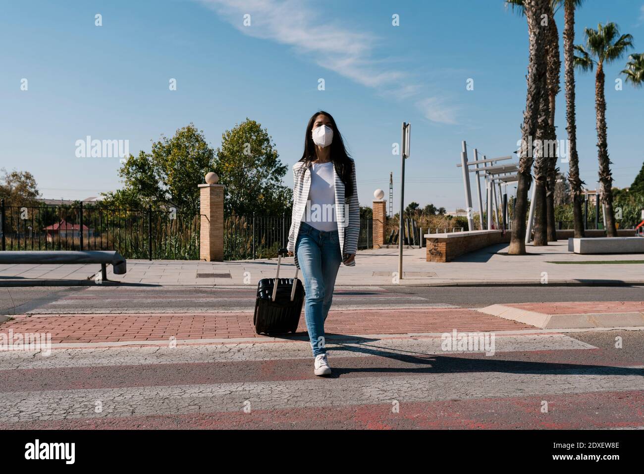Woman with luggage crossing street against blue sky on sunny day during pandemic Stock Photo