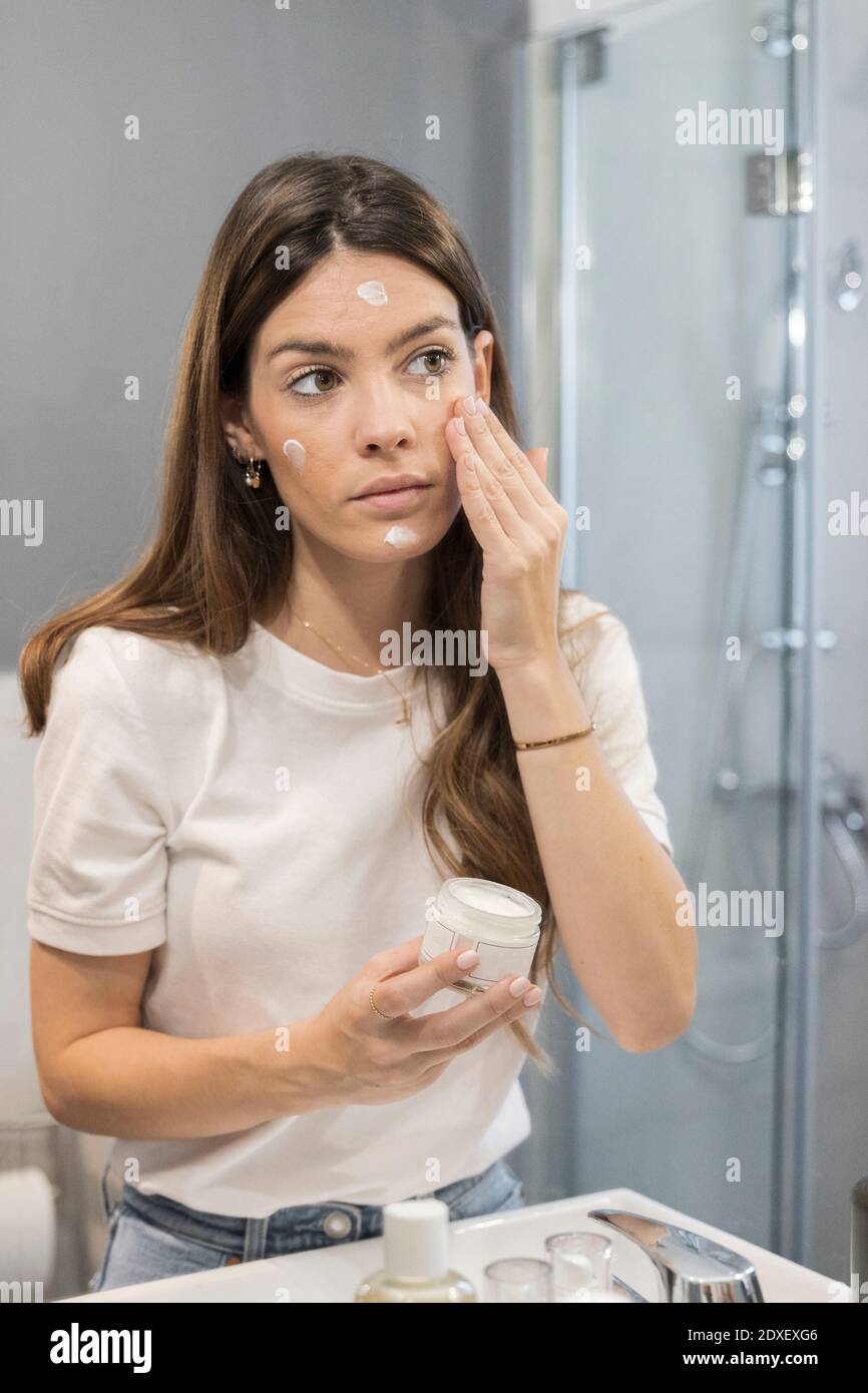 Young woman rubbing cream on face while standing in bathroom at home Stock Photo