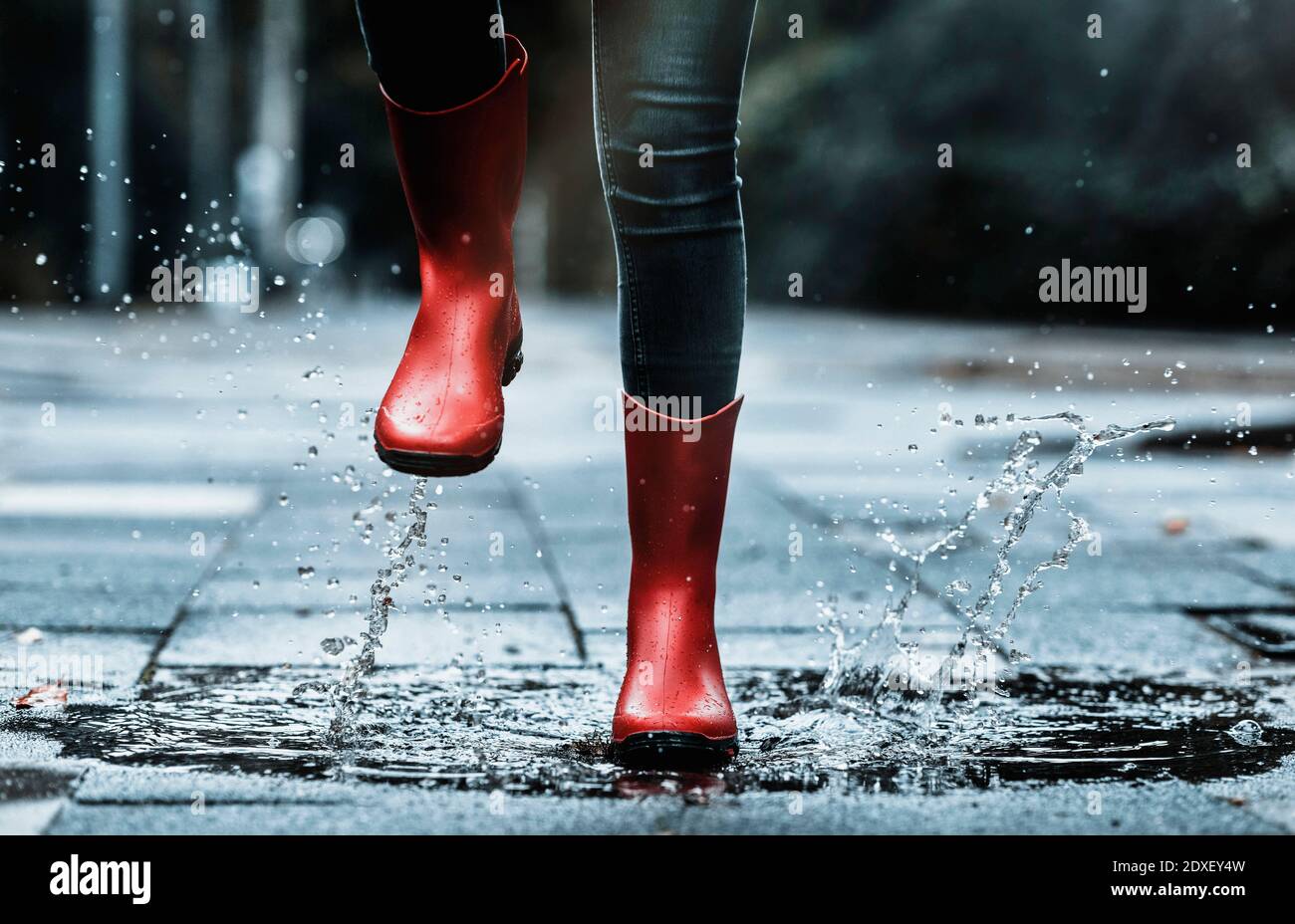 Girl wearing rubber boots splashing water while jumping in puddle on footpath Stock Photo