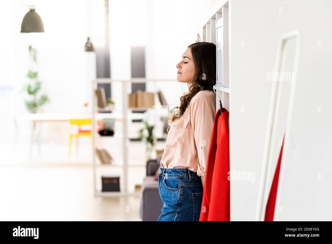 Female fashion designer with eyes closed leaning on rack at studio ...