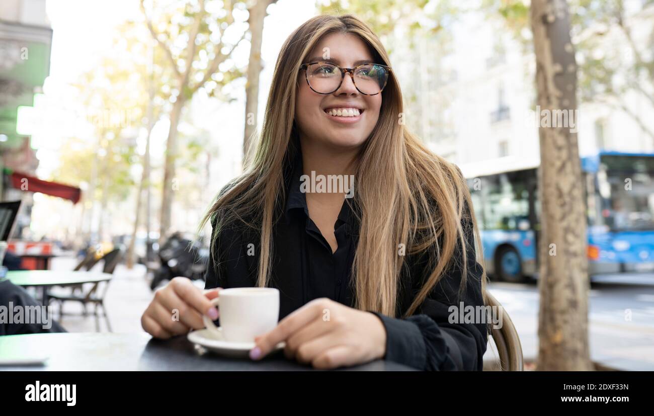 Young woman with coffee cup smiling while sitting at sidewalk cafe in city Stock Photo