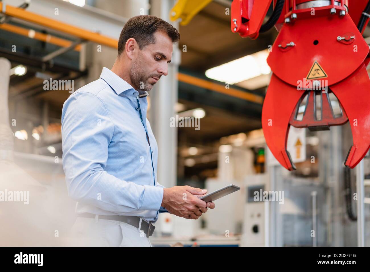 Male entrepreneur using digital tablet while standing in front of claw machinery at industry Stock Photo